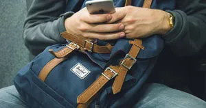 A person texting on a phone while holding a backpack.